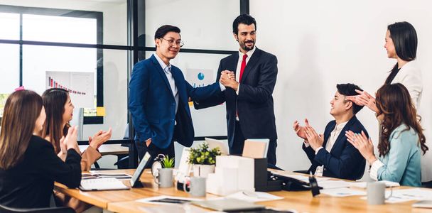Представьте себе двух азиатских деловых партнеров в элегантном костюме, удачно пожимающих друг другу руки перед группой случайных деловых людей, хлопающих в ладоши в современном офисе. - Фото, изображение