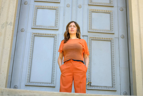 Vista de ángulo bajo de una mujer en traje naranja de colores de pie esperando con las manos en los bolsillos delante de una gran puerta de madera histórica con copyspace - Foto, imagen