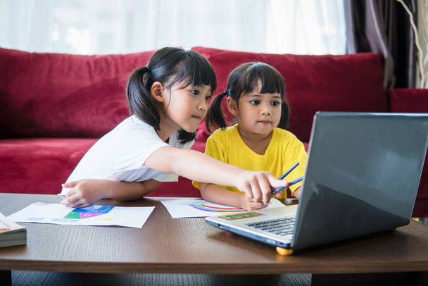 Две девочки учатся онлайн вместе с учительницей по видеосвязи. Братья и сёстры учатся дома с ноутбуком во время карантина из-за пандемии Ковида 19.. - Фото, изображение