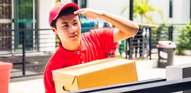 Happy delivery man in red uniform holding cardboard box standing near house of customer.courier service concept - Фото, изображение