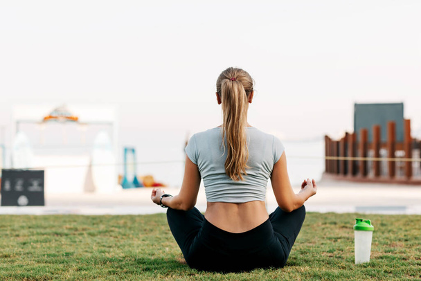 A young woman sits in a lotus position on a lawn on the Blue Waters Island in Dubai and meditates. Sports, fitness, yoga. Healthy lifestyle concept, rear view, text space - Фото, зображення