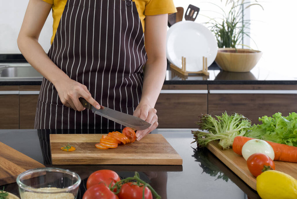 Jeune femme au foyer couper la tomate rouge en morceaux sur une planche à découper en bois. Le comptoir de cuisine plein de différents types de légumes. Ambiance matinale dans une cuisine moderne. - Photo, image
