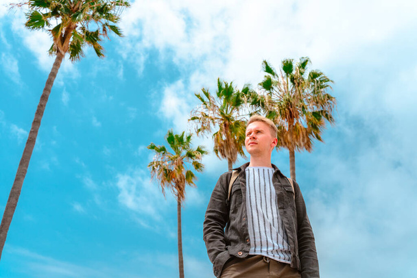 A young man in a denim jacket stands against a palm tree with a blue sky on Venice Beach in Los Angeles, photographed from below - Photo, Image