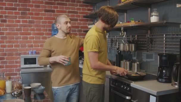 Slowmo tracking shot di uomo felice bere caffè e abbracciare il suo partner maschile cucinare la colazione in cucina Stanno ridendo e di essere affettuoso - Filmati, video