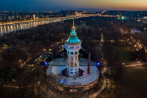 Budapest, Hungary - Aerial view of the illumimnated Margaret Island Open-Air Stage (Margitszigeti Szabadteri Szinpad) water tower at dusk with clear blue and golden sky and downtown of Budapest at background - Photo, Image