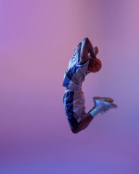 Jugador de baloncesto saltando con pelota en el estudio, fondo de neón. Baller masculino profesional en ropa deportiva juego de deporte - Foto, imagen