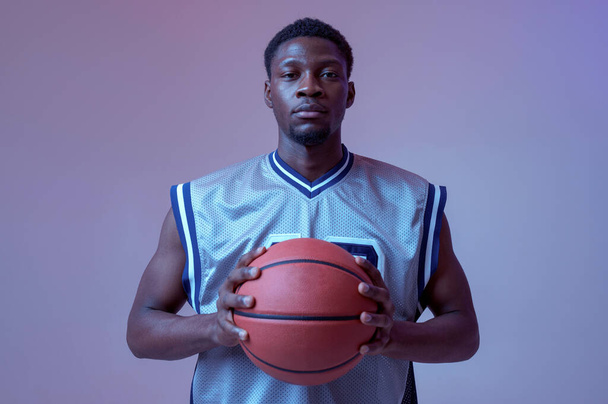 Confident basketball player poses with ball in studio, neon background. Professional male baller in sportswear playing sport game, tall sportsman - Foto, Bild
