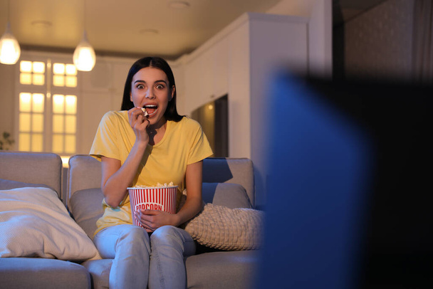 Young woman watching movie with popcorn on sofa at night - 写真・画像