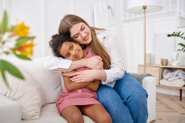 Happy mother hugs her little daughter on sofa in living room.Mom and female child leisures in their house together, good relationship, parental care - Photo, Image
