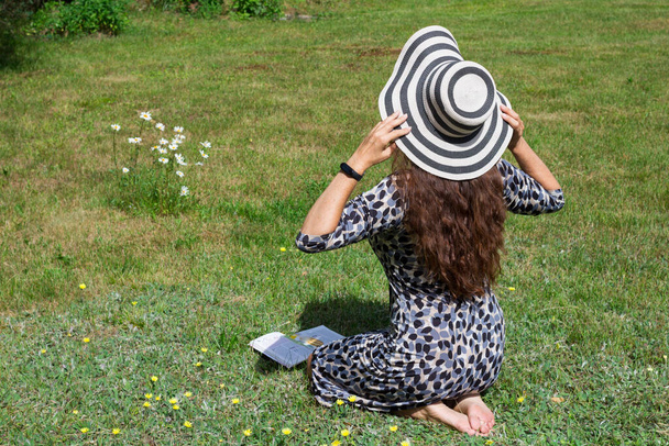 Дівчина в плямистій сукні з розкішним, хвилястим, довгим, червоним волоссям сидить на траві, тримаючи в руках смугастий капелюх
.. - Фото, зображення