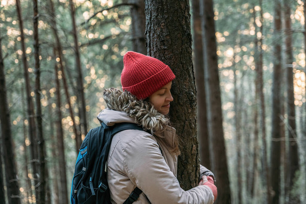 Dağ yürüyüşü sırasında soğuk ormandaki bir ağaca sarılan kırmızı şapkalı turist genç kadın. Doğaya bağlanmak, çevre kavramını korumak - Fotoğraf, Görsel