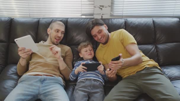 Slowmo shot of gay fathers and their cute son relaxing on couch and using gadgets - Footage, Video