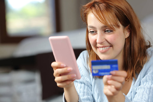 Happy woman buying online using smart phone and credit card in the bedroom at home - Photo, Image