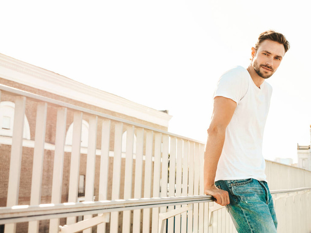 Portrait of handsome smiling  hipster lambersexual model.Stylish man dressed in white T-shirt and jeans. Fashion male posing on the street background near fence at the beach at sunset - Zdjęcie, obraz