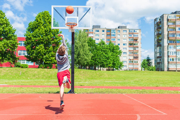 Vonzó fiatal fiú lövés labdát a karika a játszótéren.aranyos fiatal fiú kosarazik a játszótéren a nyári meleg napon. - Fotó, kép