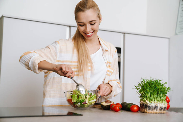 Joyful blonde woman smiling while preparing fresh salad at home kitchen - Foto, afbeelding