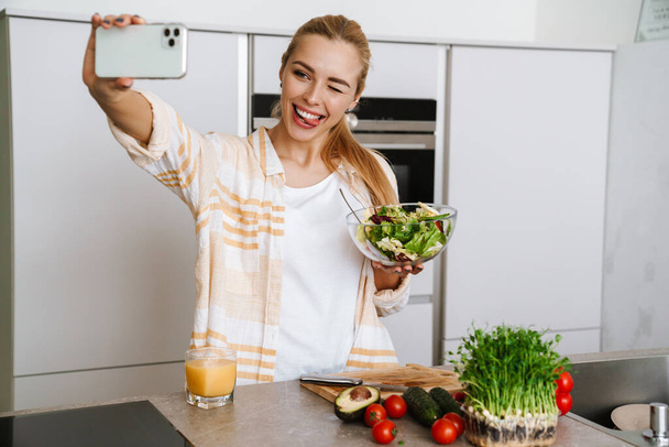 Joyful blonde woman taking selfie on cellphone with fresh salad at home kitchen - Foto, afbeelding