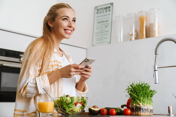Portrait of a happy attractive woman using mobile phone while making a healthy salad in the kitchen - Fotografie, Obrázek