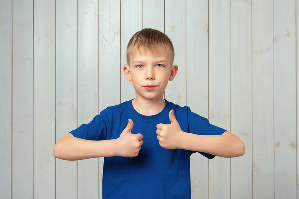 Jóképű, fiatal fiú kék pólóban, mosolyogva és boldogan mutogatja a hüvelykujját, dicséret a választásért. Stúdió lövés, világos fa háttér - Fotó, kép