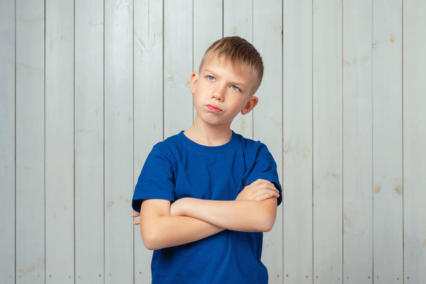 Portrait of moody displeased preteen boy in blue t shirt, crossing arms on her chest, looking aside. Studio shot, light wooden background - Photo, image