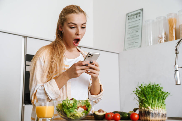 Retrato de una mujer feliz emocionada usando el teléfono móvil mientras hace una ensalada saludable en la cocina - Foto, Imagen