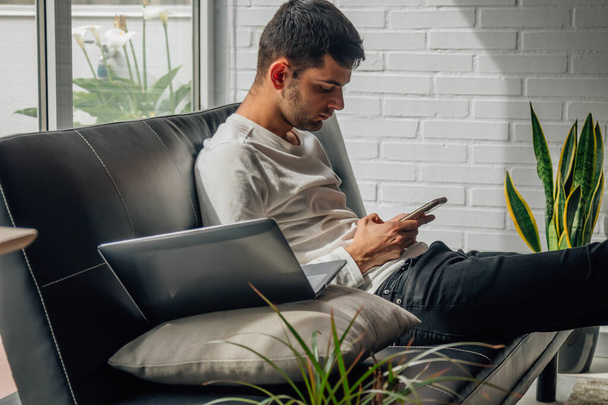 mladý muž s mobilním telefonem doma sedí na pohovce - Fotografie, Obrázek