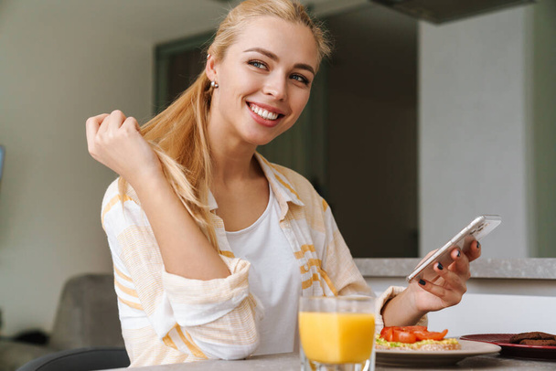 Happy beautiful young woman using mobile phone while having tasty healthy breakfast in a kitchen at home - Photo, Image