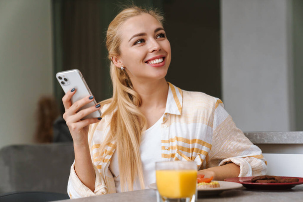 Happy beautiful young woman using mobile phone while having tasty healthy breakfast in a kitchen at home - Foto, Imagem