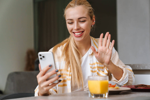 Feliz hermosa mujer joven usando el teléfono móvil para una videollamada mientras desayuna sabroso y saludable en una cocina en casa - Foto, Imagen