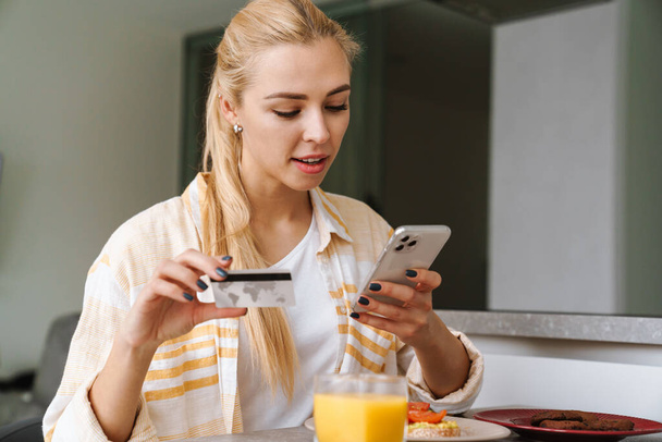 Pleased woman using cellphone and credit card while having breakfast at home kitchen - Fotó, kép