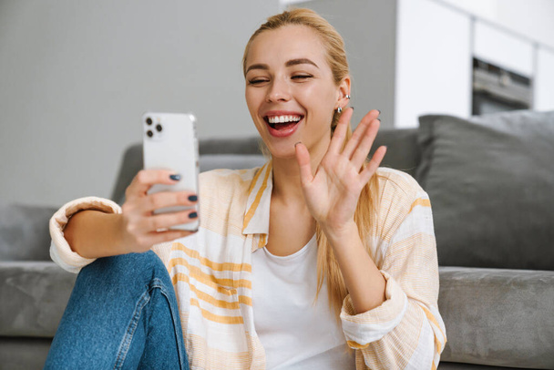 Happy blonde woman waving hand while taking selfie on cellphone at home - Foto, imagen