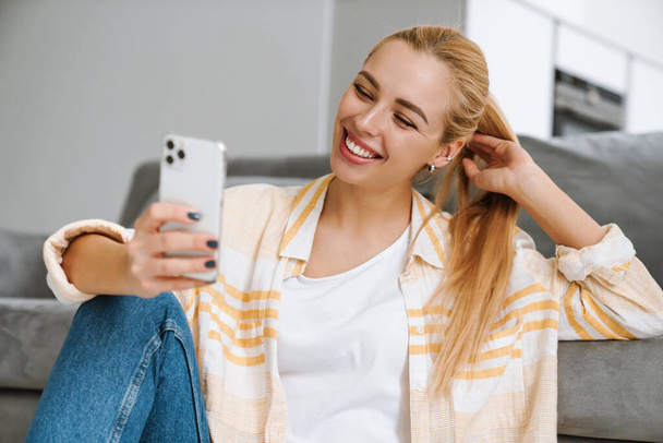 Happy blonde woman smiling and using cellphone while sitting on floor at home - Φωτογραφία, εικόνα