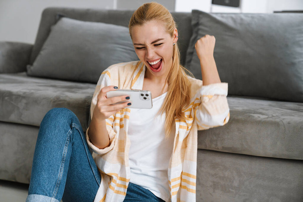 Cheerful excited young girl using mobile phone while sitting on a floor at the couch at home - Foto, imagen