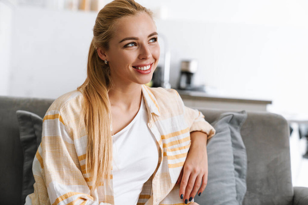 Happy blonde woman smiling while sitting on couch at home - Fotó, kép