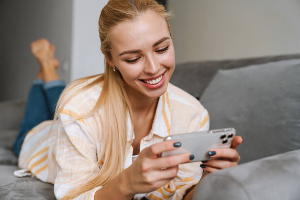 Happy blonde woman smiling and using cellphone while lying on couch at home - Fotografie, Obrázek