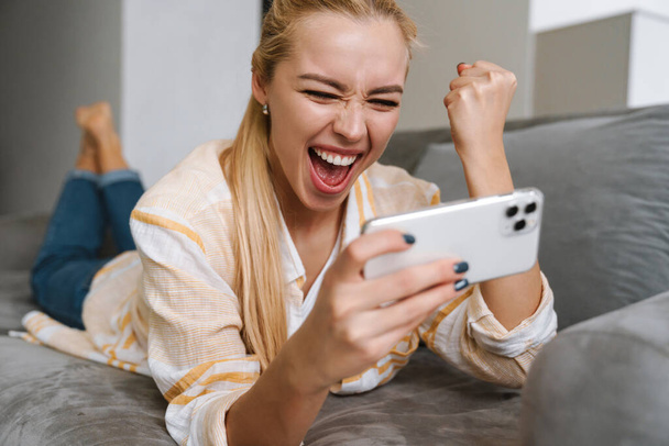 Excited woman making winner gesture while playing online game on cellphone at home - Fotó, kép