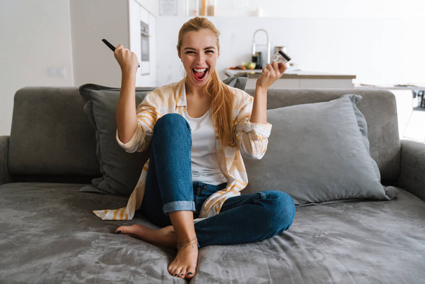 Excited woman holding credit card while watching tv with remote control at home - Foto, immagini