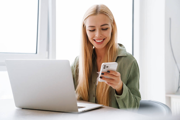 Happy smiling attractive young woman with laptop computer using mobile phone at home office - Фото, изображение