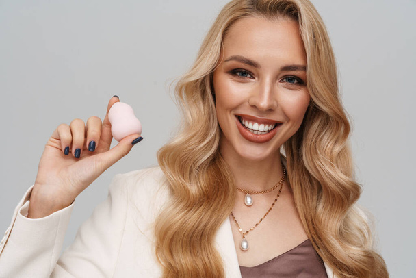 Happy gorgeous woman smiling while posing with cosmetic sponge isolated over grey background - Φωτογραφία, εικόνα