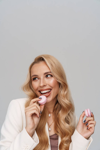 Beautiful happy girl in jacket smiling while eating macaroons isolated over grey background - Foto, afbeelding