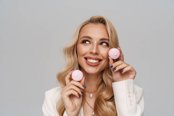 Beautiful happy girl in jacket smiling while posing with macaroons isolated over grey background - Foto, afbeelding