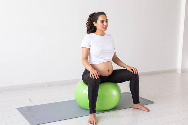 Een jonge zwangere vrouw doet ontspanningsoefening met een fitnessbal terwijl ze op een mat zit. Kopieerruimte - Foto, afbeelding