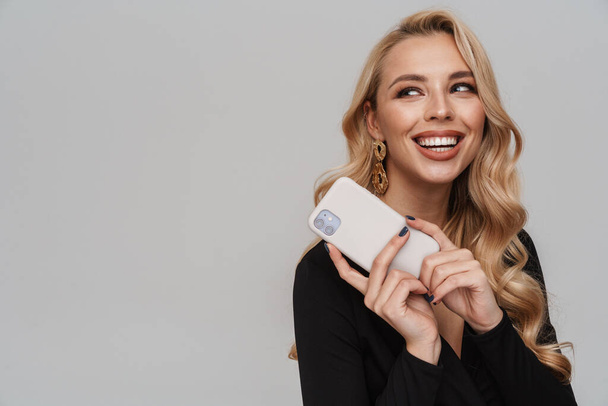 Happy girl in luxury earrings smiling while posing with cellphone isolated over grey background - Φωτογραφία, εικόνα