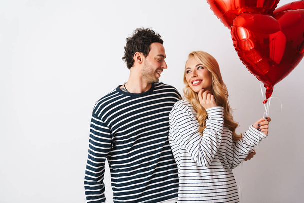 Glückliches romantisches Paar lächelt, während es mit Herzballons vor weißem Hintergrund posiert - Foto, Bild