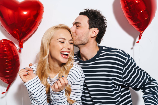Joyful couple kissing and making fun while posing with heart balloons isolated over white background - Fotoğraf, Görsel