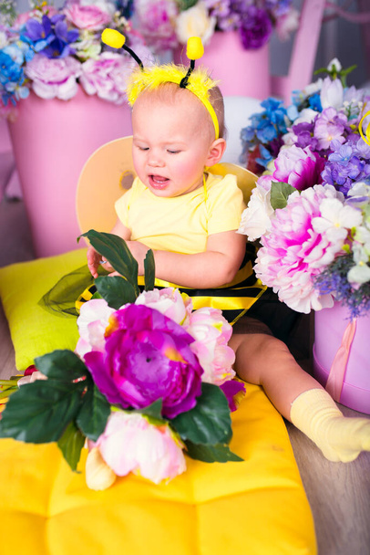 Een babymeisje in een kostuum van een bij zit op een geel kussen tussen heldere bloemen. Haar mond is open en ze kijkt naar beneden.. - Foto, afbeelding