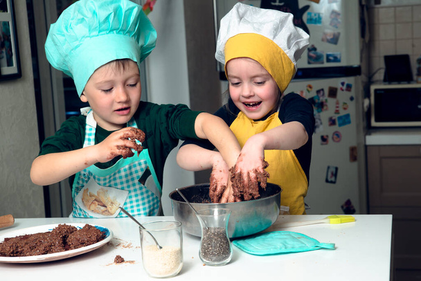 Siblings in cooking hats and aprons are buisy with kneading brown daugh in a bowl, both are happy, hands smudged. Concept of children cooking. - Photo, Image