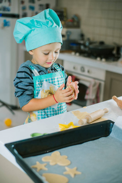 Egy cuki fiú szakácsjelmezben, aki tésztát gyúrt. Süti vágó, serpenyő, sodrófa és tészta fekszik az asztalon. - Fotó, kép