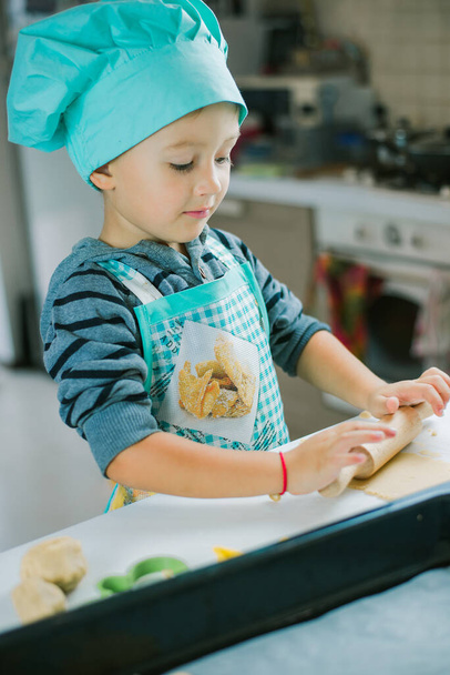 Милый мальчик в костюме повара разворачивает выпечку с видом на кухню на заднем плане. На столе лежат ножи, сковорода и тесто. - Фото, изображение