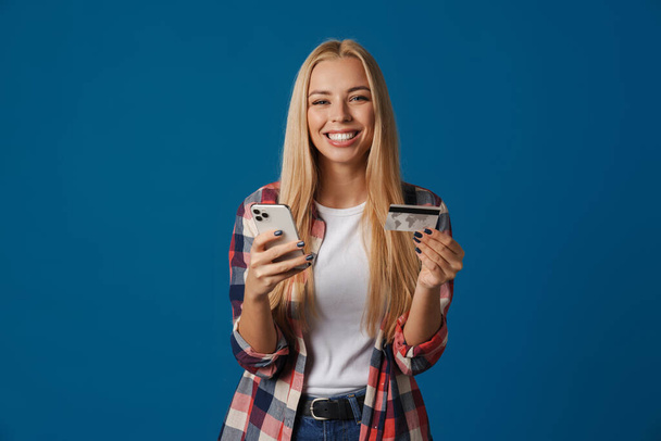 Loira feliz mulher usando celular e mostrando cartão de crédito isolado sobre fundo azul - Foto, Imagem